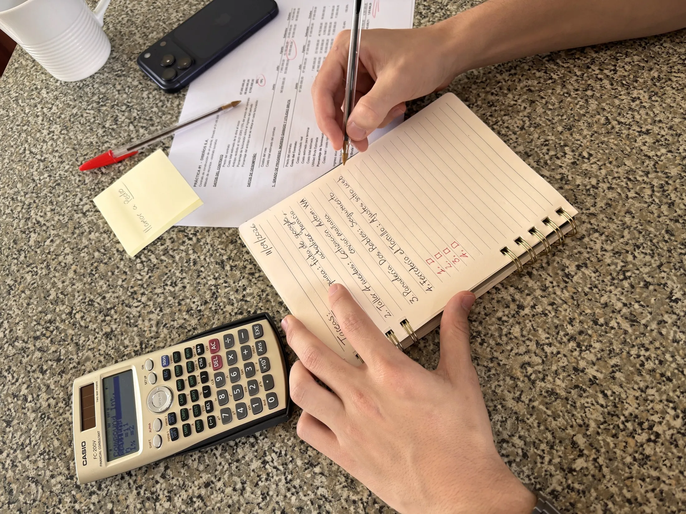 Manos revisando hojas de pedidos y anotando prioridades en una libreta, junto a una calculadora y un teléfono sobre una mesa de trabajo.